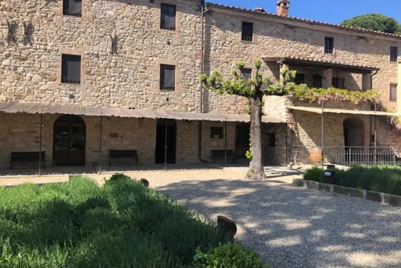 A stone building faces a gravel courtyard with a leafy tree.