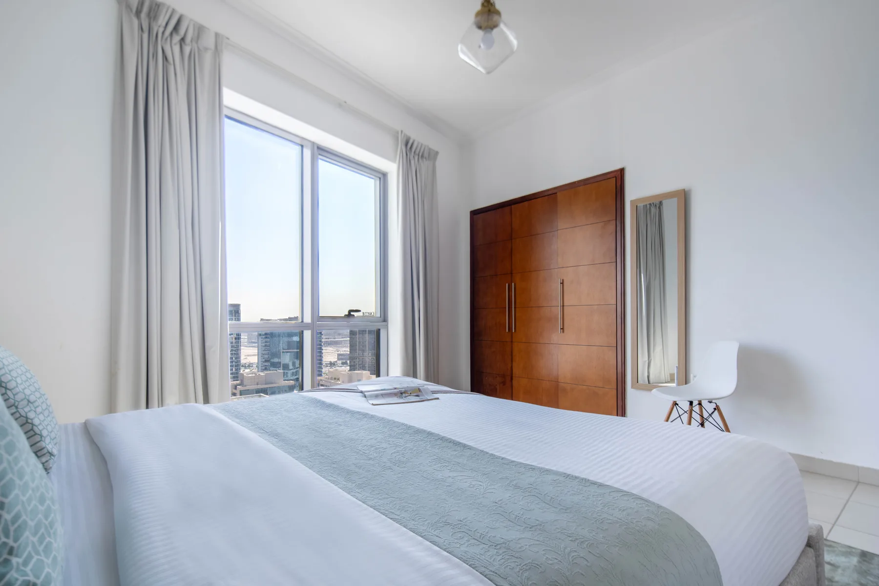 Un dormitorio luminoso con una cama grande junto a una ventana con vistas a edificios de la ciudad.