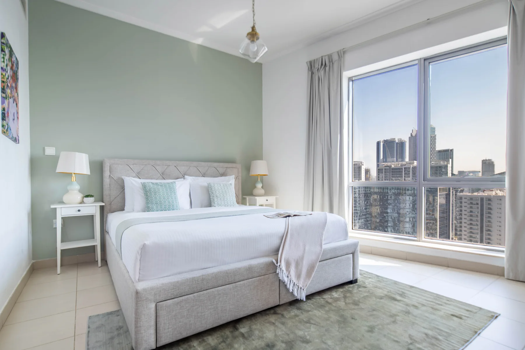 Un dormitorio luminoso tiene una cama bien arreglada junto a grandes ventanas con vistas a edificios de la ciudad.