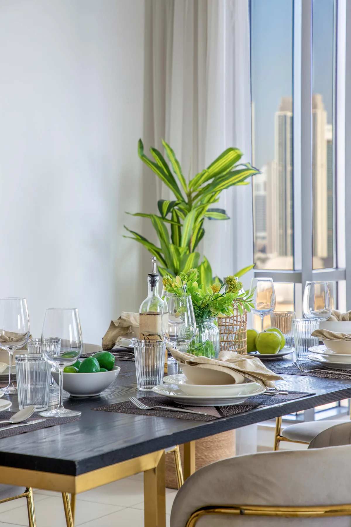 Una mesa de comedor está puesta con platos y vasos junto a una planta frondosa y ventanas altas.