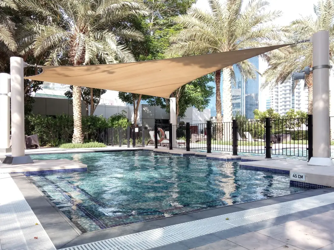Una piscina al aire libre está bajo una lona de sombra, con palmeras cerca.