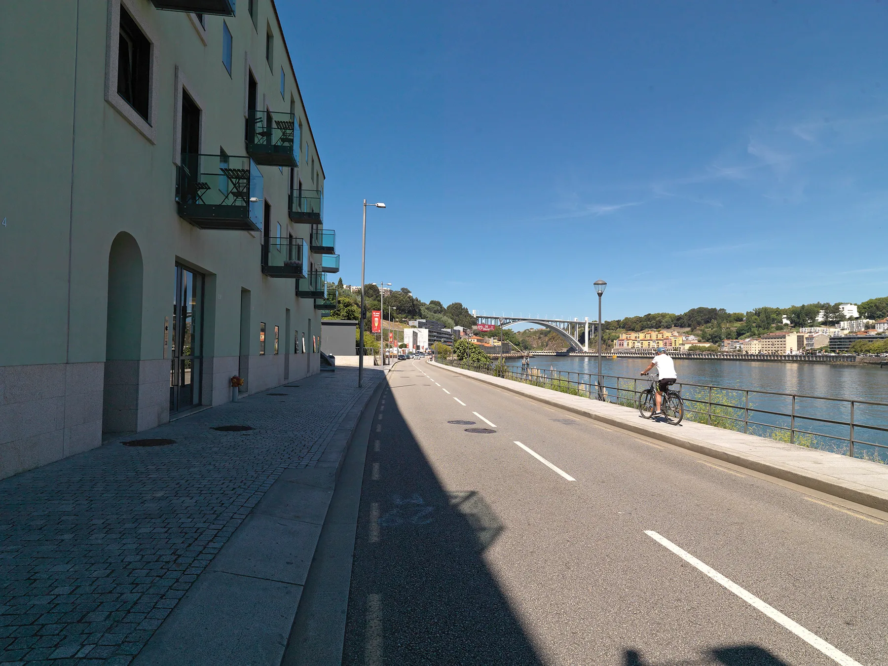 A riverside road runs beside buildings, with a cyclist and a bridge in the distance.