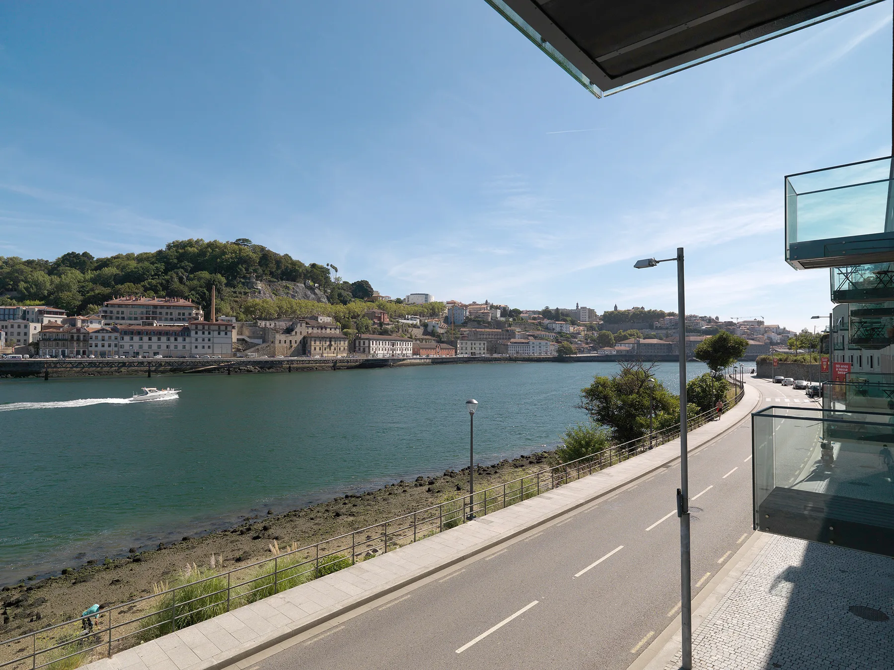 A riverfront road curves along the water, with boats and hillside buildings in the distance.