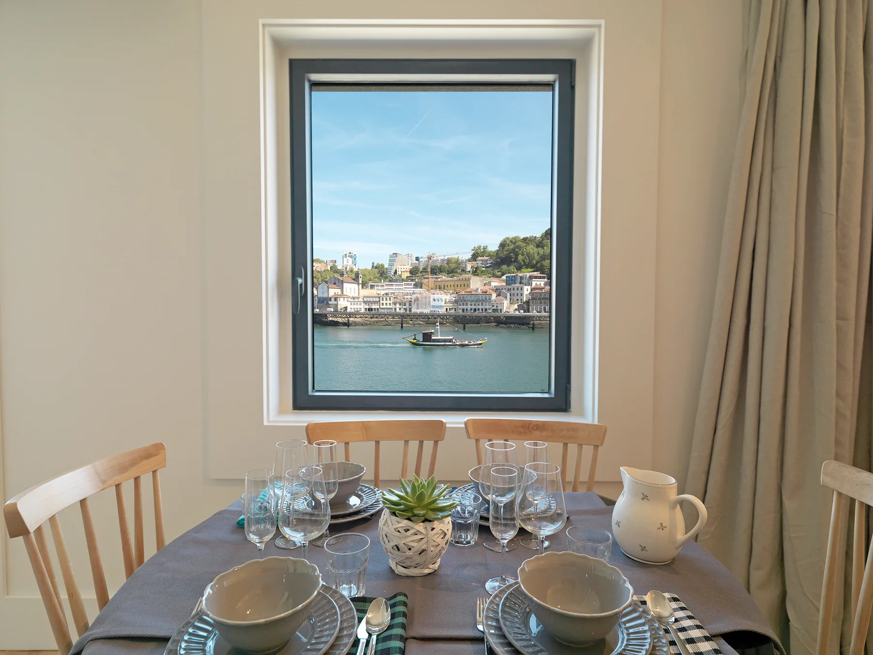A dining table by a window overlooks a river and the buildings across it.