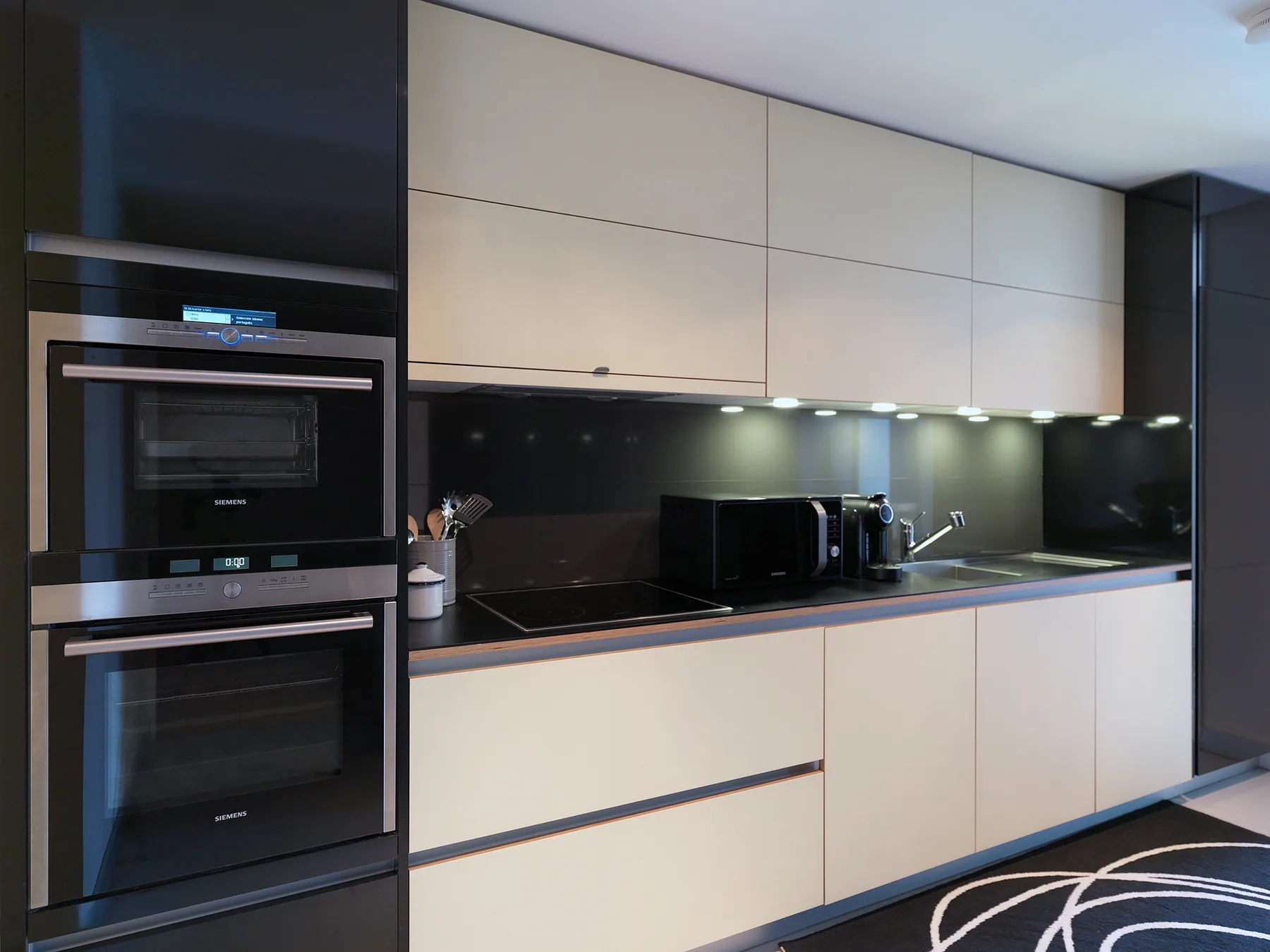 A modern kitchen with built-in ovens, pale cabinets, and a dark countertop.