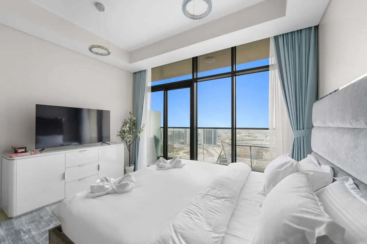 A bright bedroom with white bedding and large windows overlooking the city.