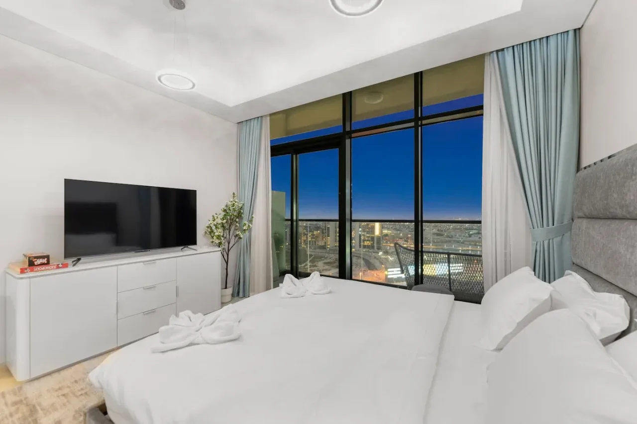 A bedroom with white bedding and floor-to-ceiling windows overlooking city lights.