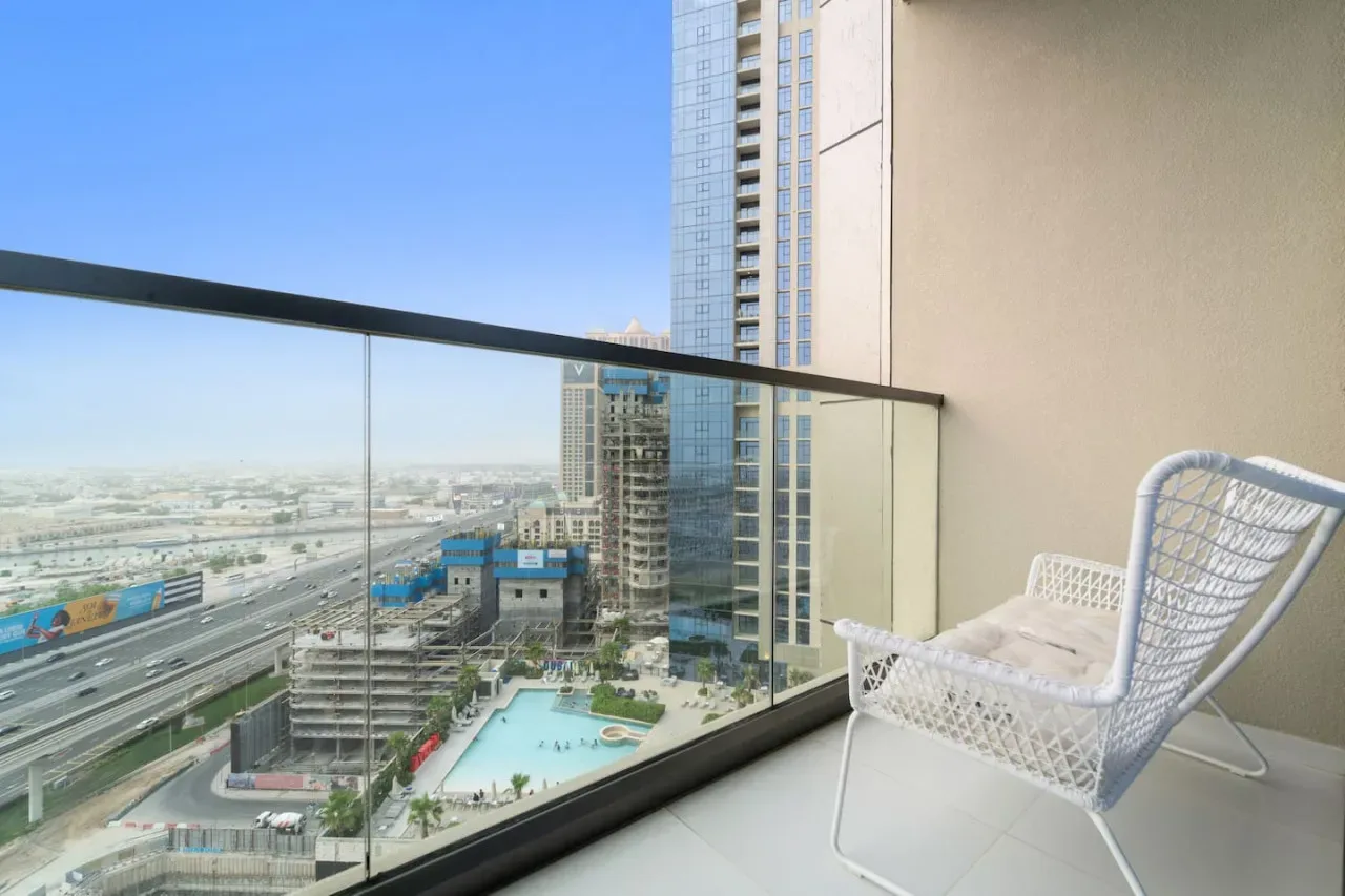 A balcony with a white woven chair overlooks high-rise buildings and a pool.