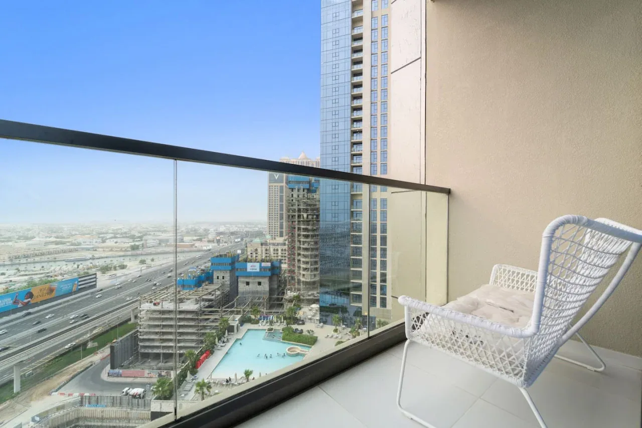 A balcony with a white woven chair overlooks high-rise buildings and a swimming pool.