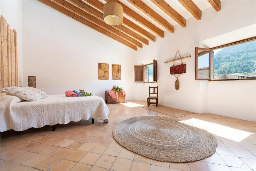 A bright bedroom with a bed, exposed wooden ceiling beams, and windows overlooking green hills.