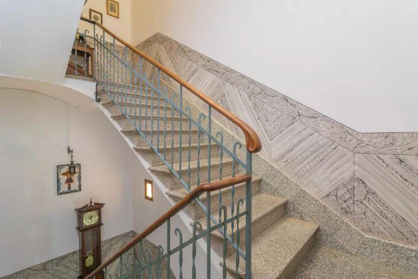 A curved indoor staircase with ornate railings rises beside a patterned stone wall.