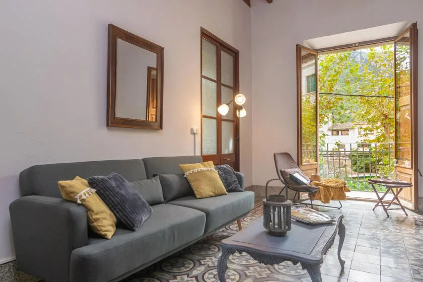A living room with a gray sofa and open doors to a leafy balcony.