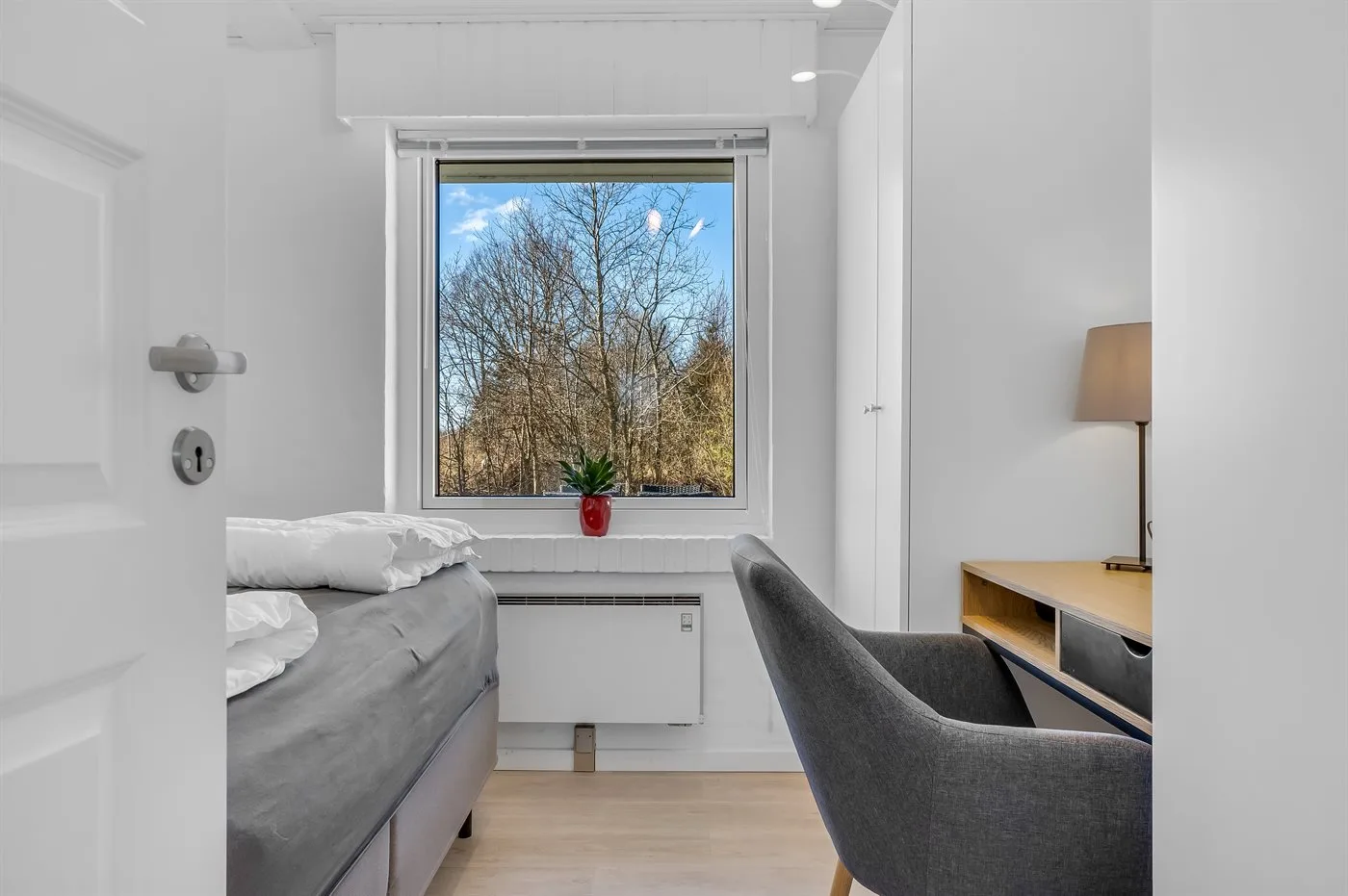 Un dormitorio luminoso con cama, escritorio y una ventana con vistas a los árboles.