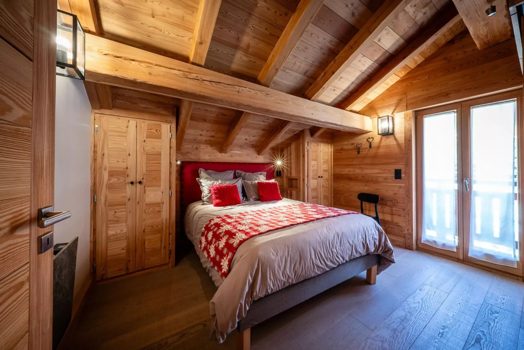A wooden bedroom with a bed, exposed ceiling beams, and tall windows.