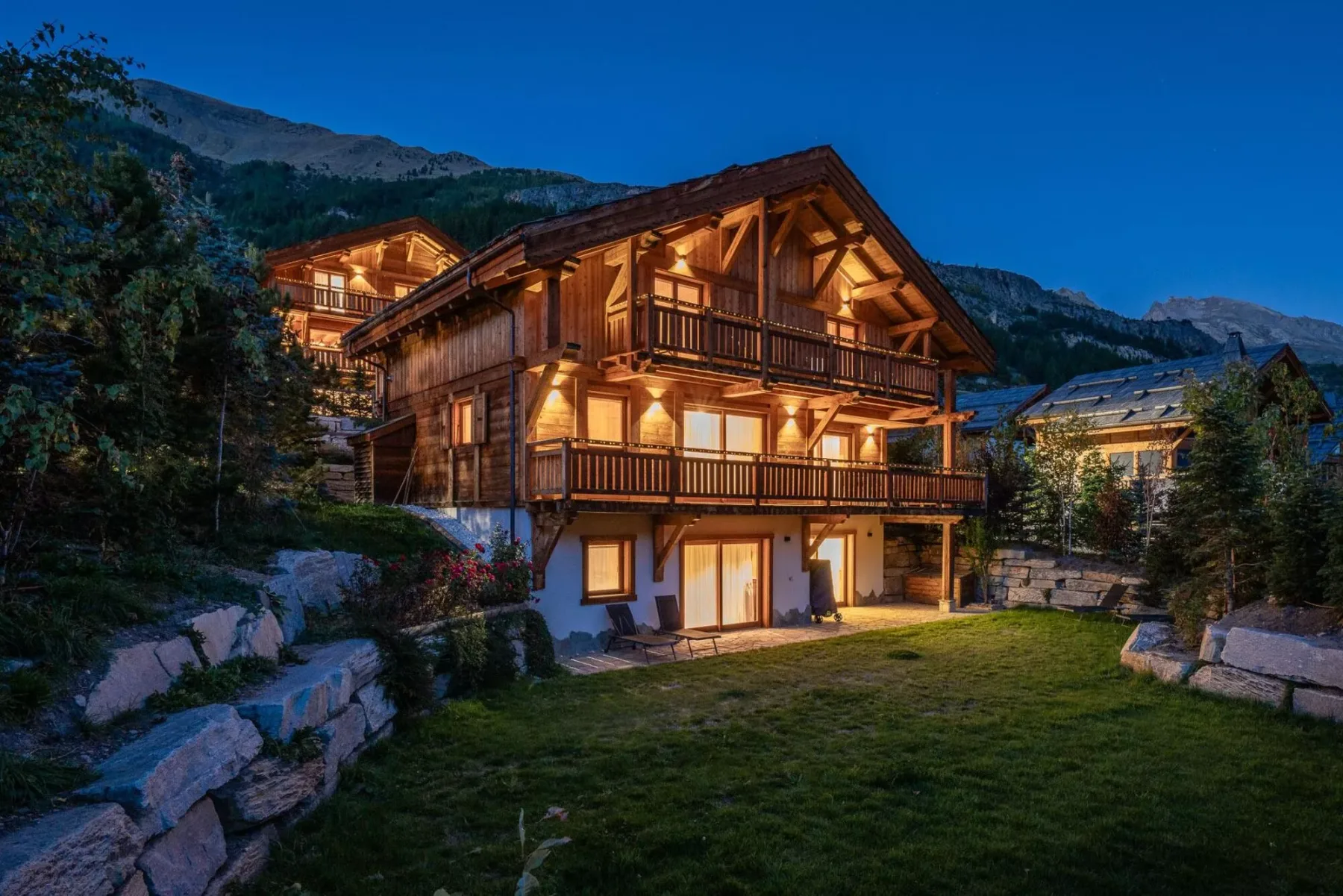 A warmly lit chalet stands beside a grassy yard beneath a blue evening sky.
