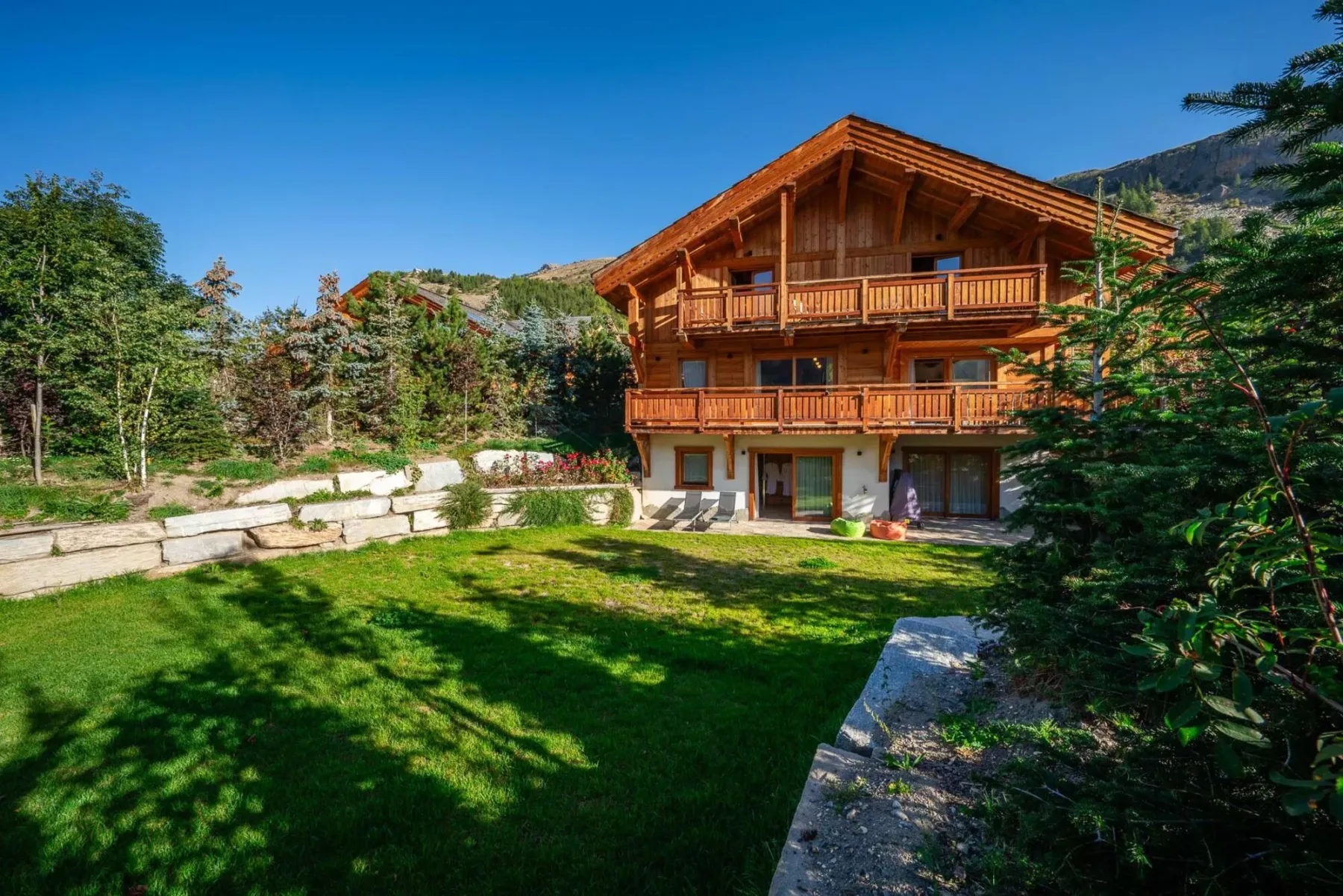 A wooden chalet stands beside a green lawn, with trees and hills in the background.