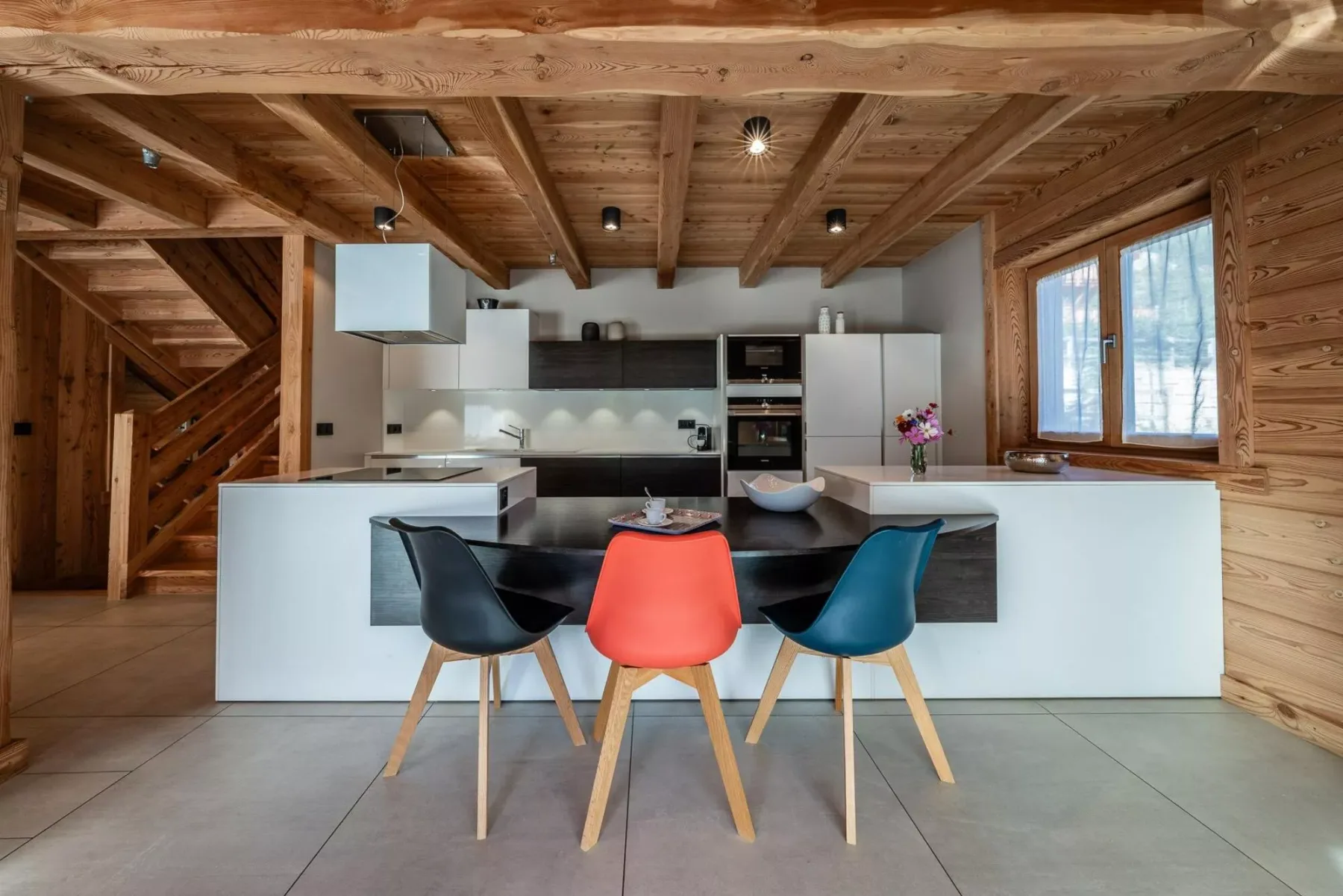 A modern kitchen with a central island and colorful seating beneath exposed wooden beams.