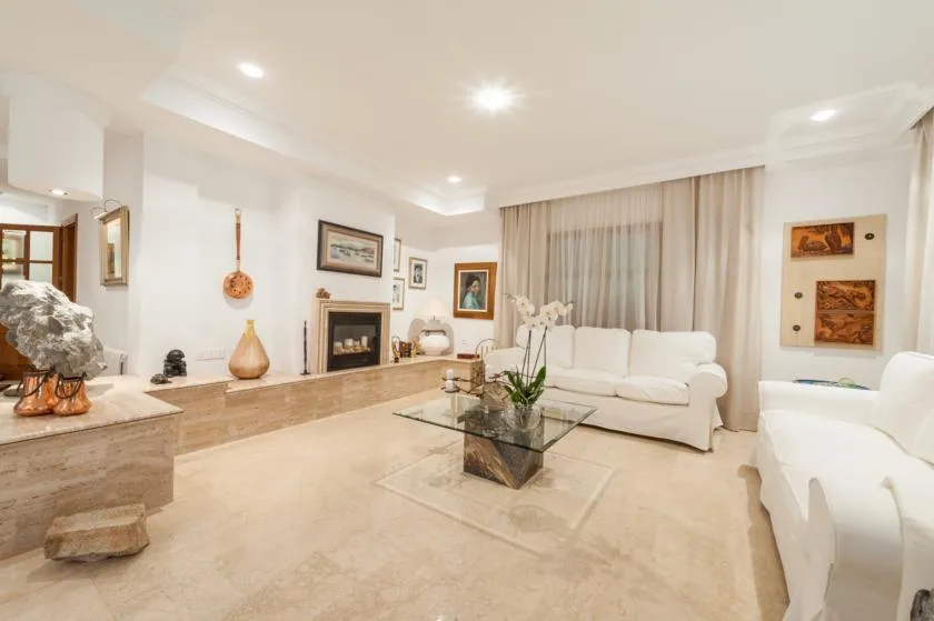 A bright living room with white seating, a glass coffee table, and a fireplace.
