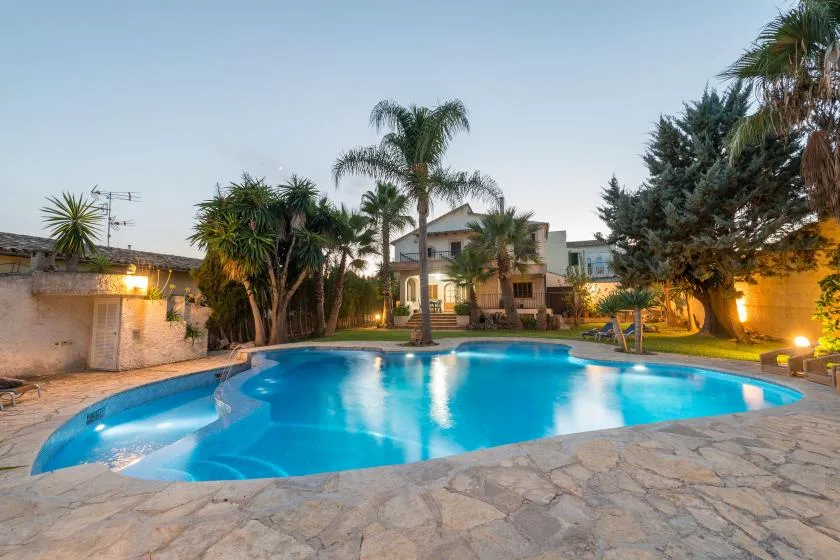 A lit swimming pool sits among palm trees and landscaped grounds.