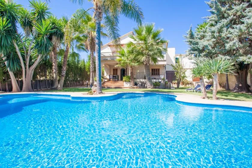 A large outdoor swimming pool sits in front of a house surrounded by palm trees.