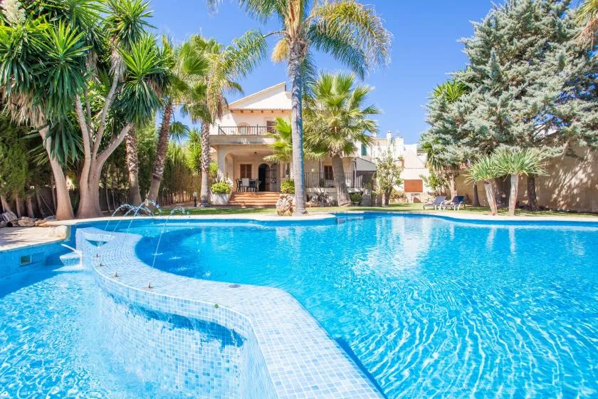 A swimming pool in front of a house, surrounded by palm trees.