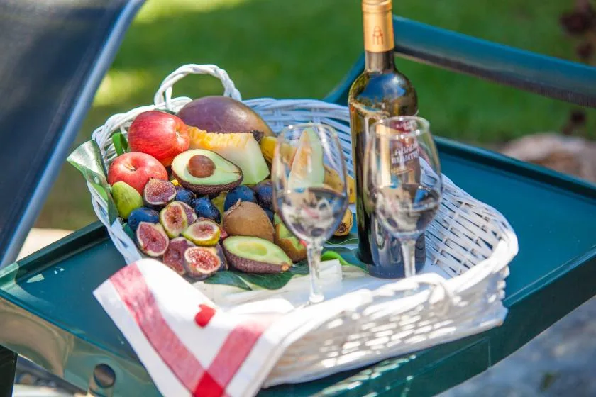 A fruit basket, wine bottle, and glasses sit on an outdoor table.