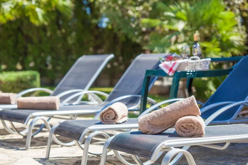 Outdoor poolside loungers with rolled towels sit beside a table in a leafy setting.