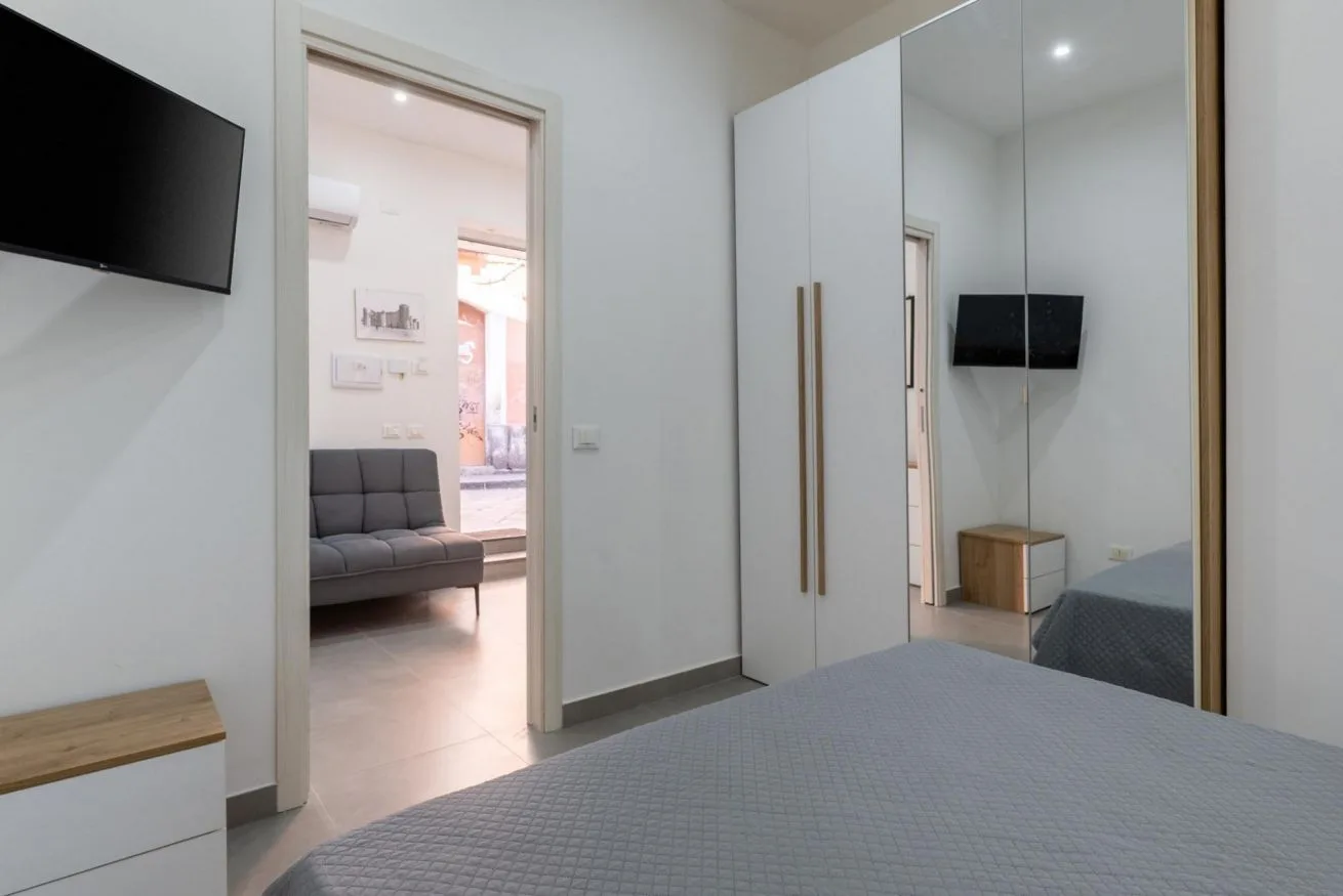 A bedroom with a bed, mirrored wardrobe, and doorway opening onto a sitting area.