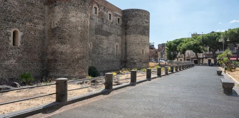 A paved outdoor walkway runs beside a large stone fortification and trees.