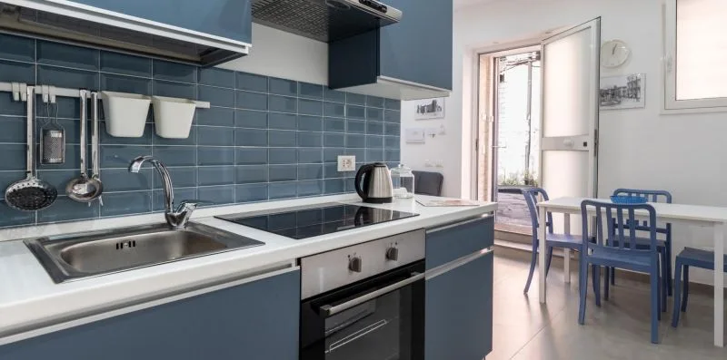 A kitchen with a sink and cooktop beside a dining table and blue chairs.