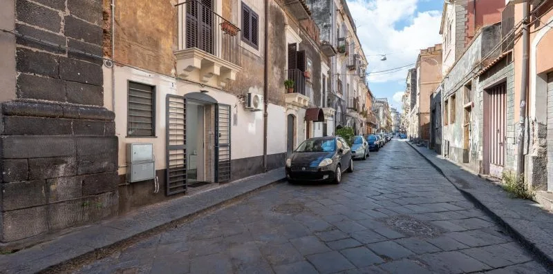 A narrow paved street runs between closely spaced buildings, with cars parked along the sides.