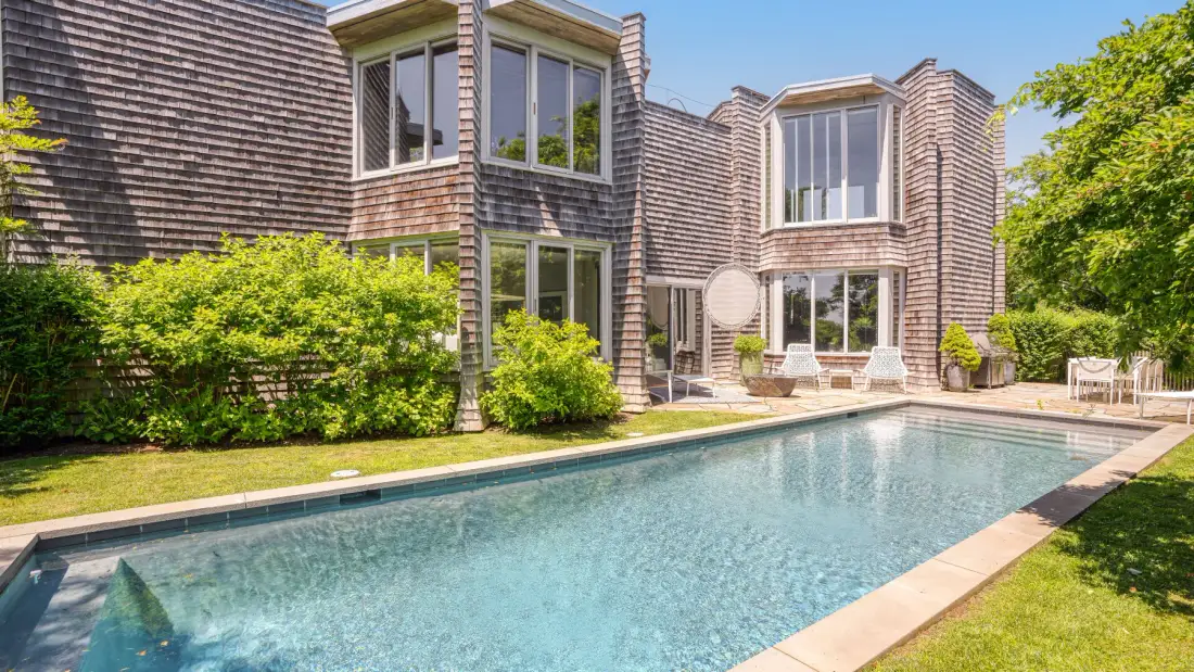 A swimming pool sits beside a shingle-clad house and green lawn.