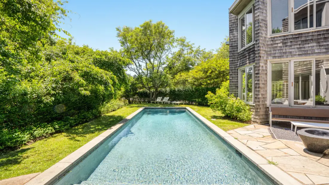 An outdoor swimming pool sits beside a house and leafy garden.