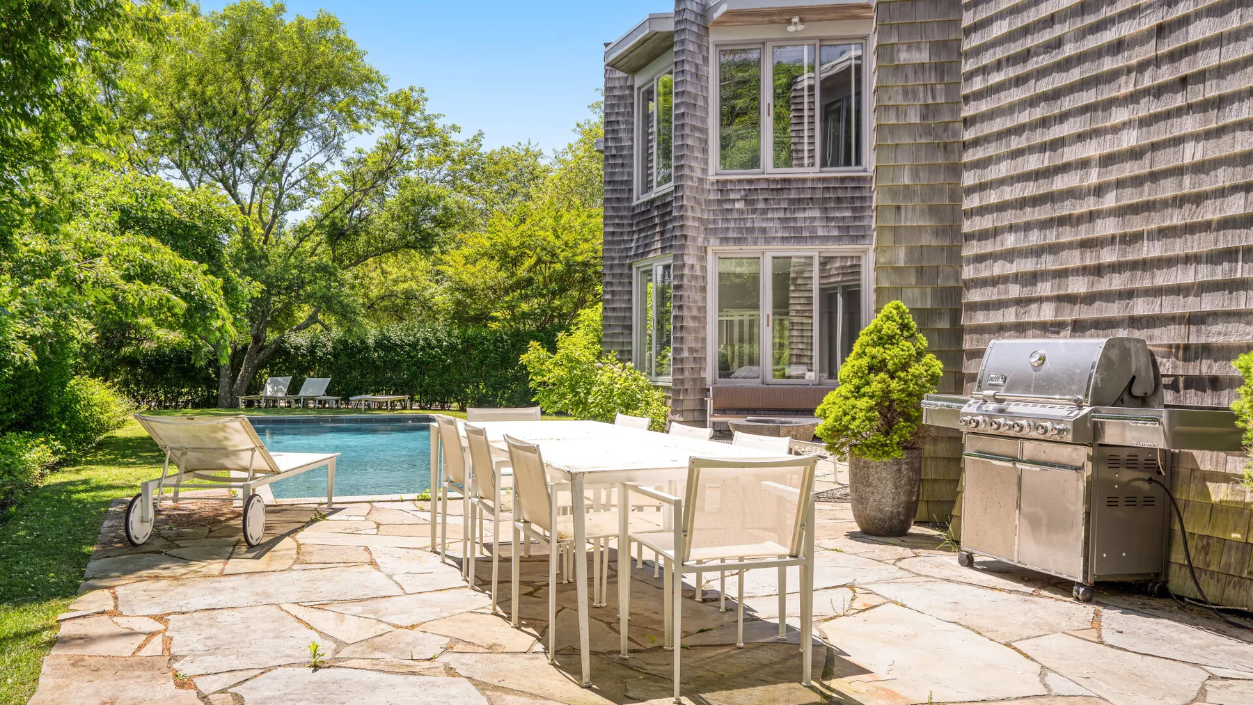 A stone patio with outdoor dining seating, a swimming pool, and a grill.