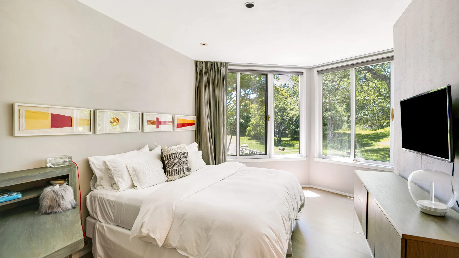 A bright bedroom with a bed beside large windows overlooking greenery.