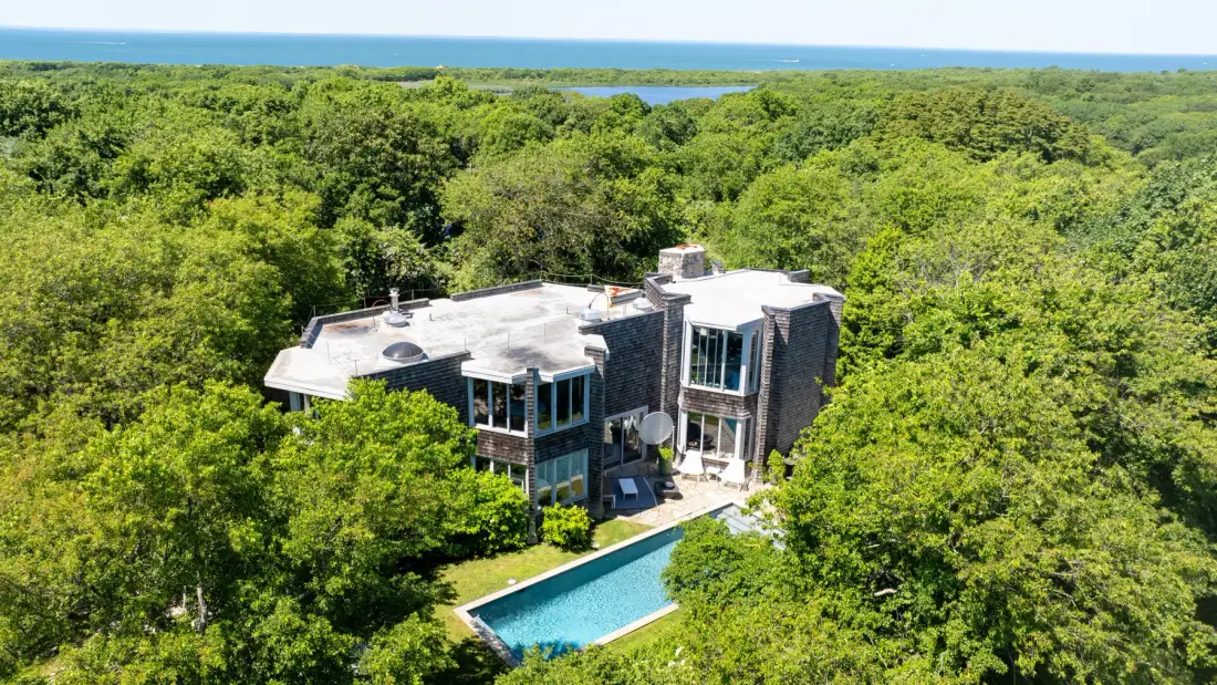 A modern house and pool sit among dense green trees, with water visible in the distance.