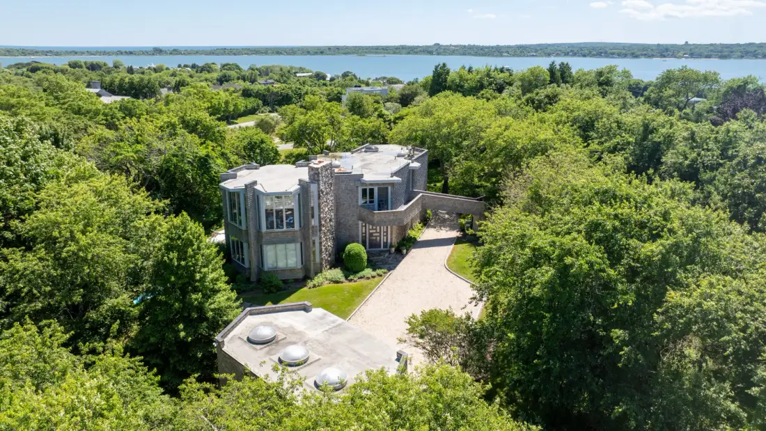 A modern house sits among dense green trees, with a paved driveway leading to it.