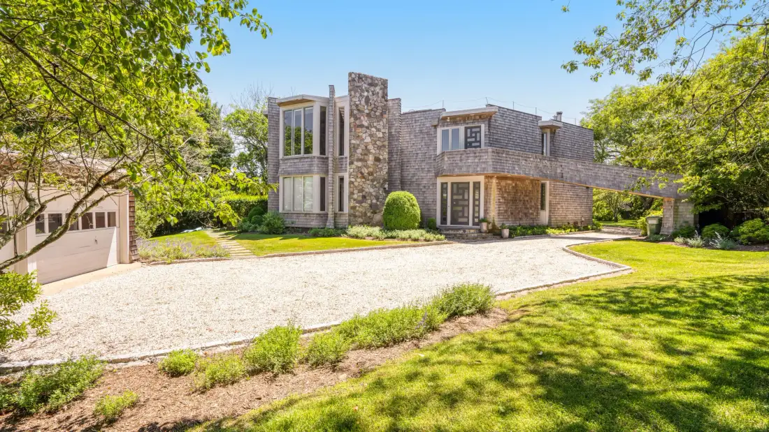 A modern stone-clad house stands beside a broad gravel driveway and green lawn.