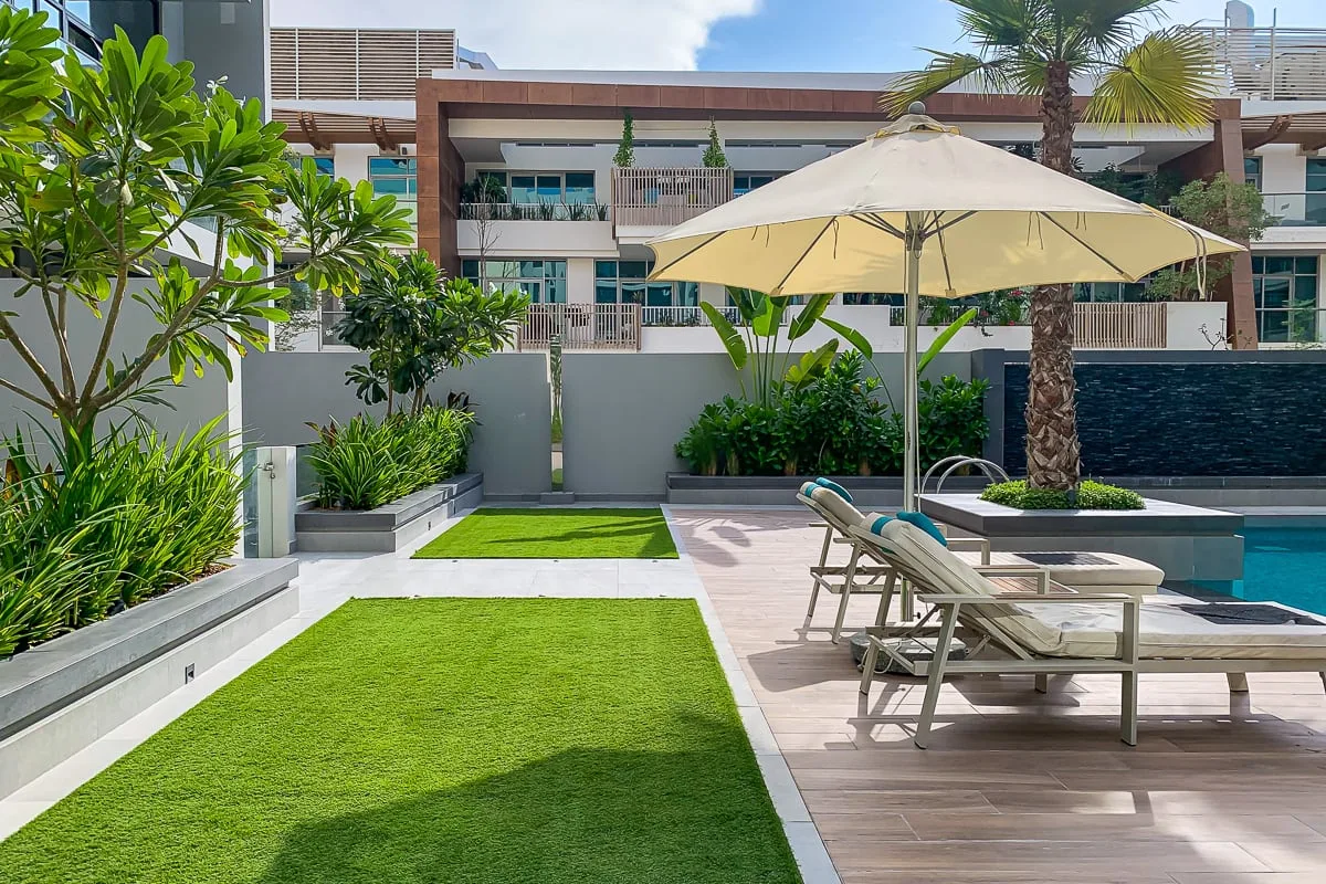 Un patio ajardinado junto a la piscina, con asientos, una sombrilla y césped verde.