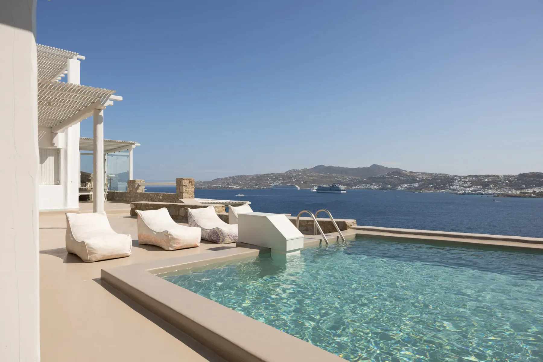 A swimming pool beside the sea, with lounge seating and distant hills.