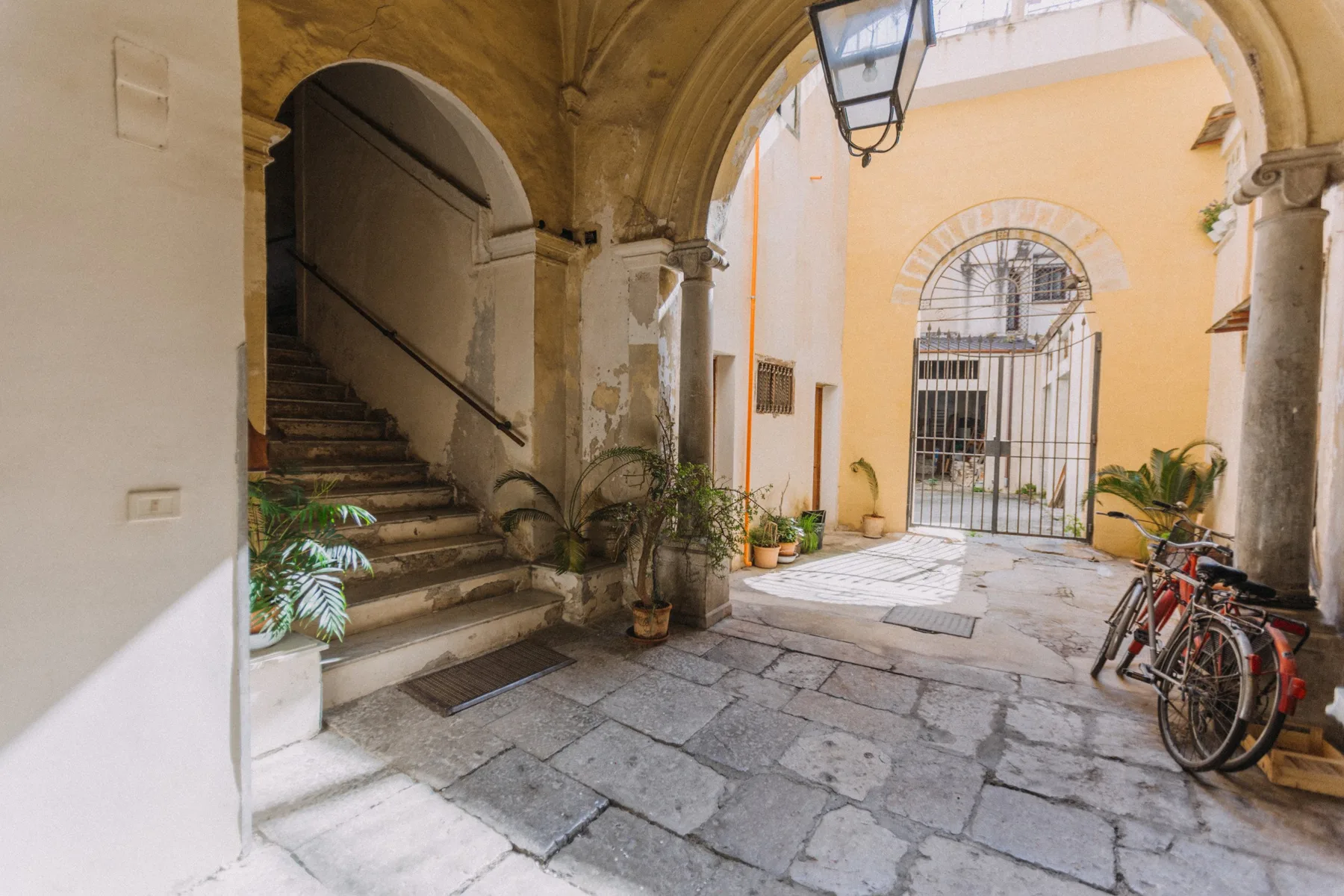Un pasaje de patio de piedra cuenta con una escalera arqueada, plantas en macetas y bicicletas.