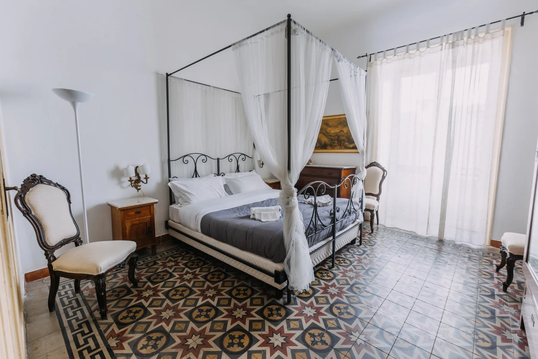 Un dormitorio con cama con dosel, suelo de baldosas estampadas y ventanas altas con cortinas translúcidas.