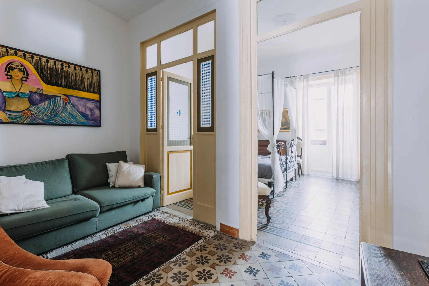 Una zona de estar con asientos verdes y una puerta que da a un dormitorio luminoso.