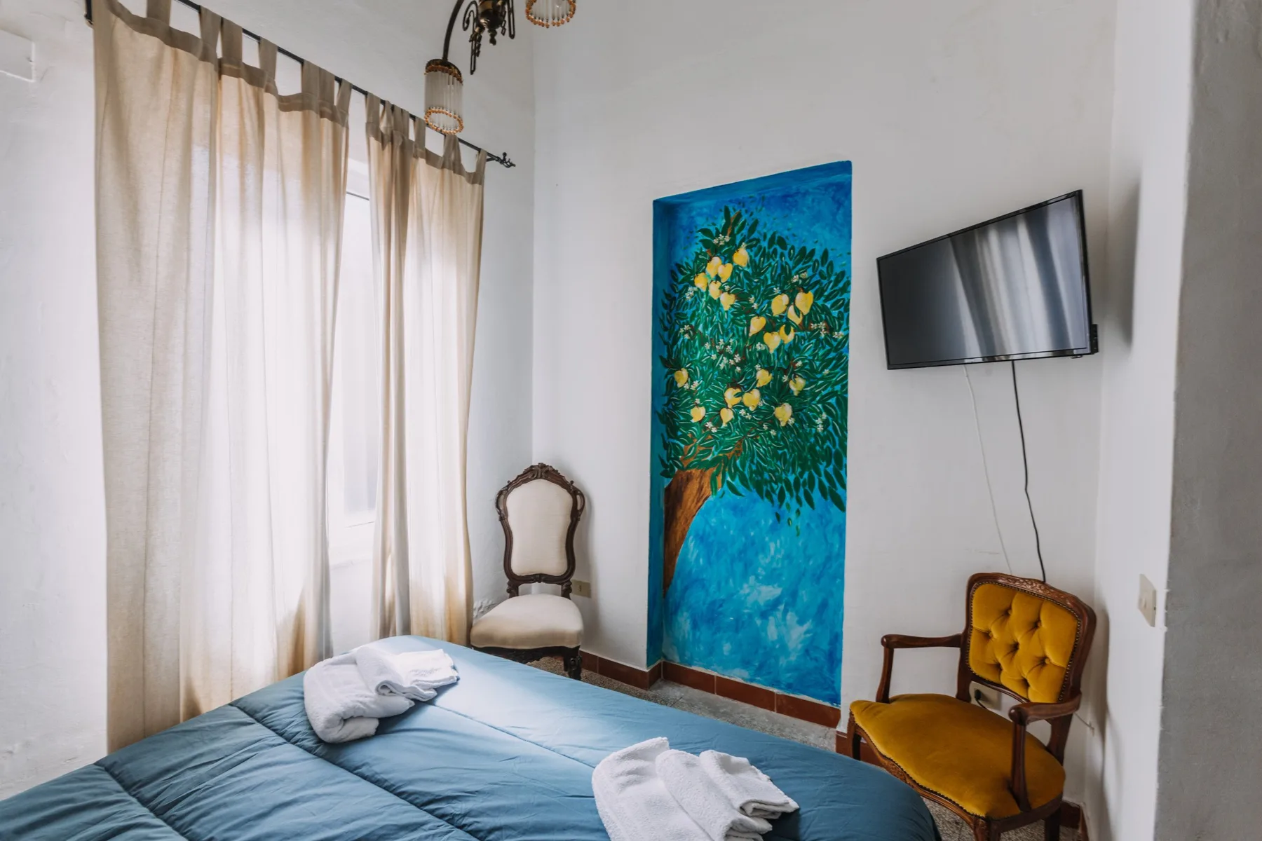 A bedroom with a blue bed, wall-mounted television, and colorful wall painting.