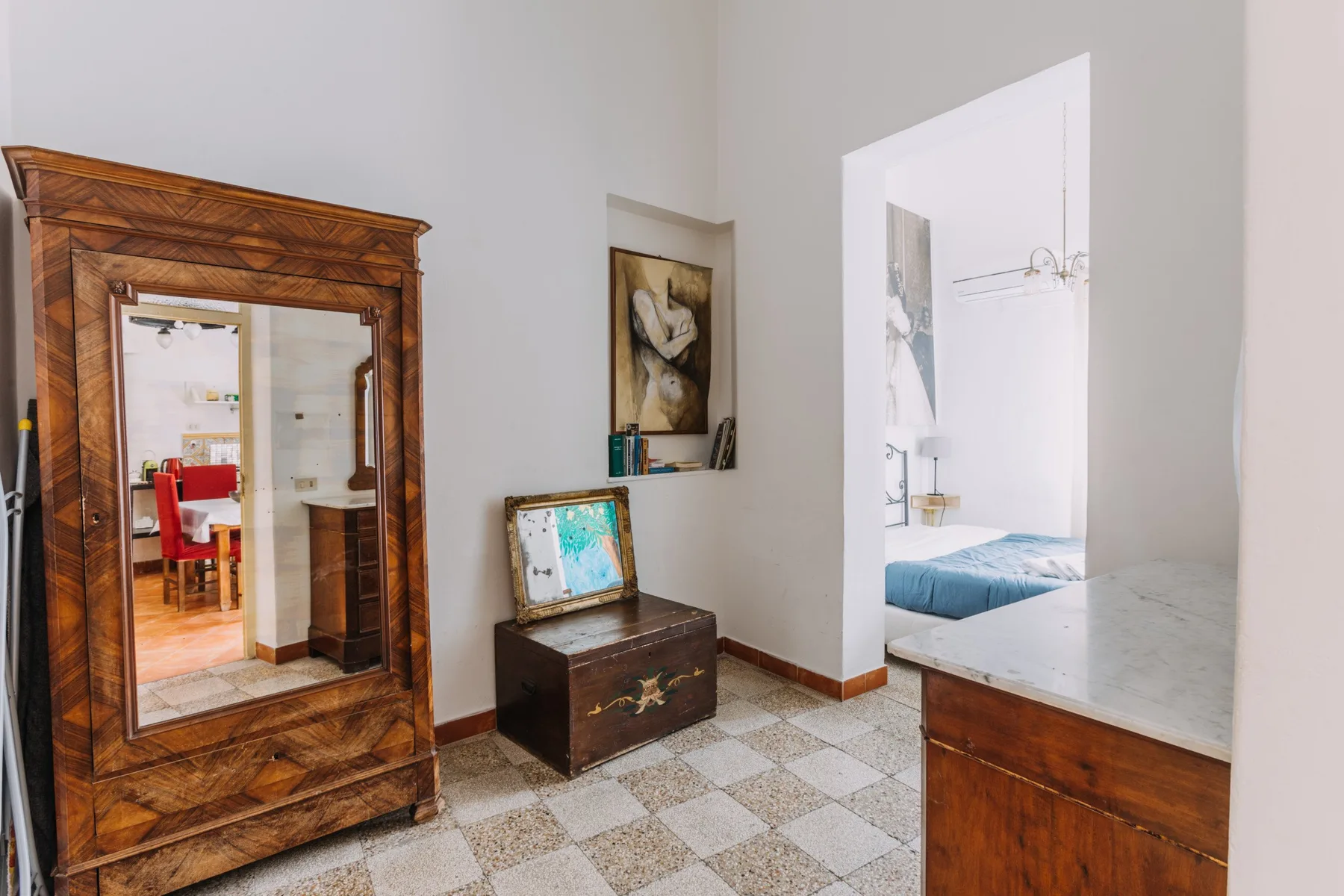Una puerta da a un dormitorio junto a un armario y un baúl de madera sobre un suelo de baldosas.