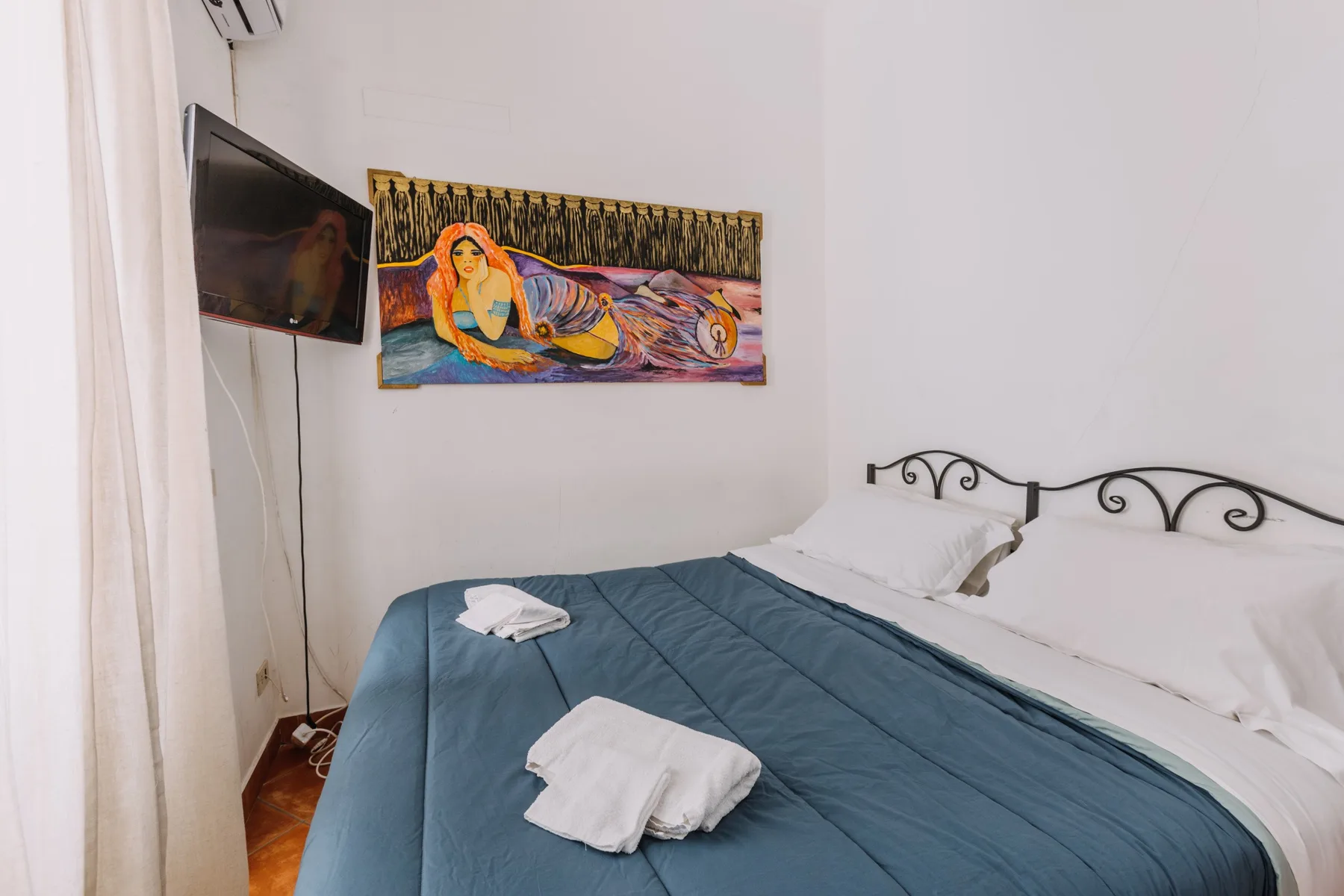 A bedroom with a blue bedspread, wall-mounted television, and colorful artwork.