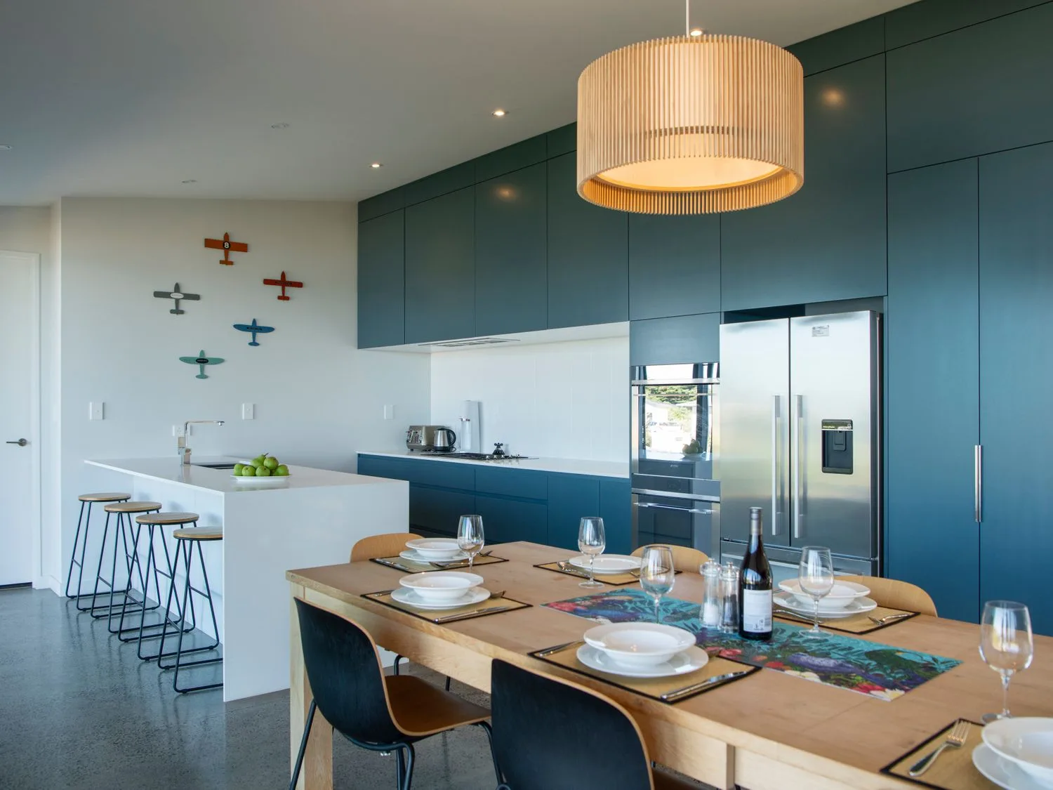 Una cocina con muebles azules, una mesa de comedor puesta con platos, vasos y cubiertos, y una isla blanca con taburetes de bar en Lightning Ridge Views - Waimarama Holiday Home, en Waimarama