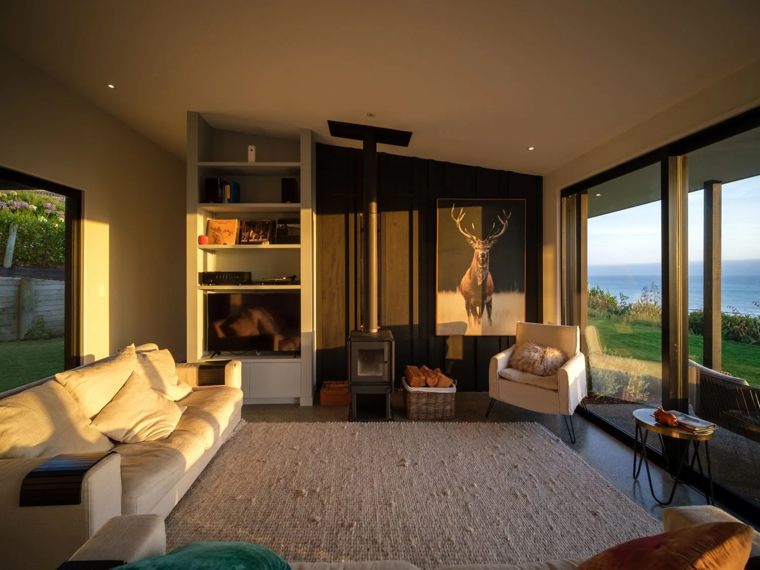 A living room with seating, a wood stove, built-in shelving, and windows overlooking greenery and the ocean.