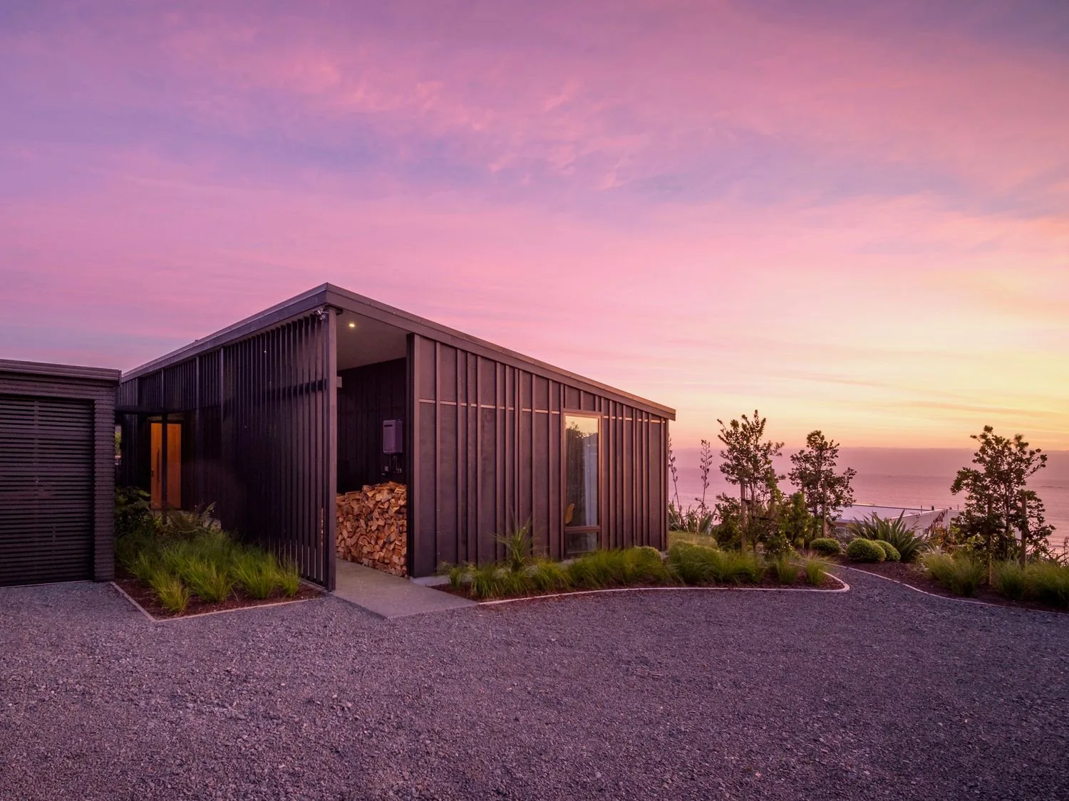 A modern house with black vertical siding a gravel driveway and greenery under a purple and orange sunset sky at Lightning Ridge Views Waimarama