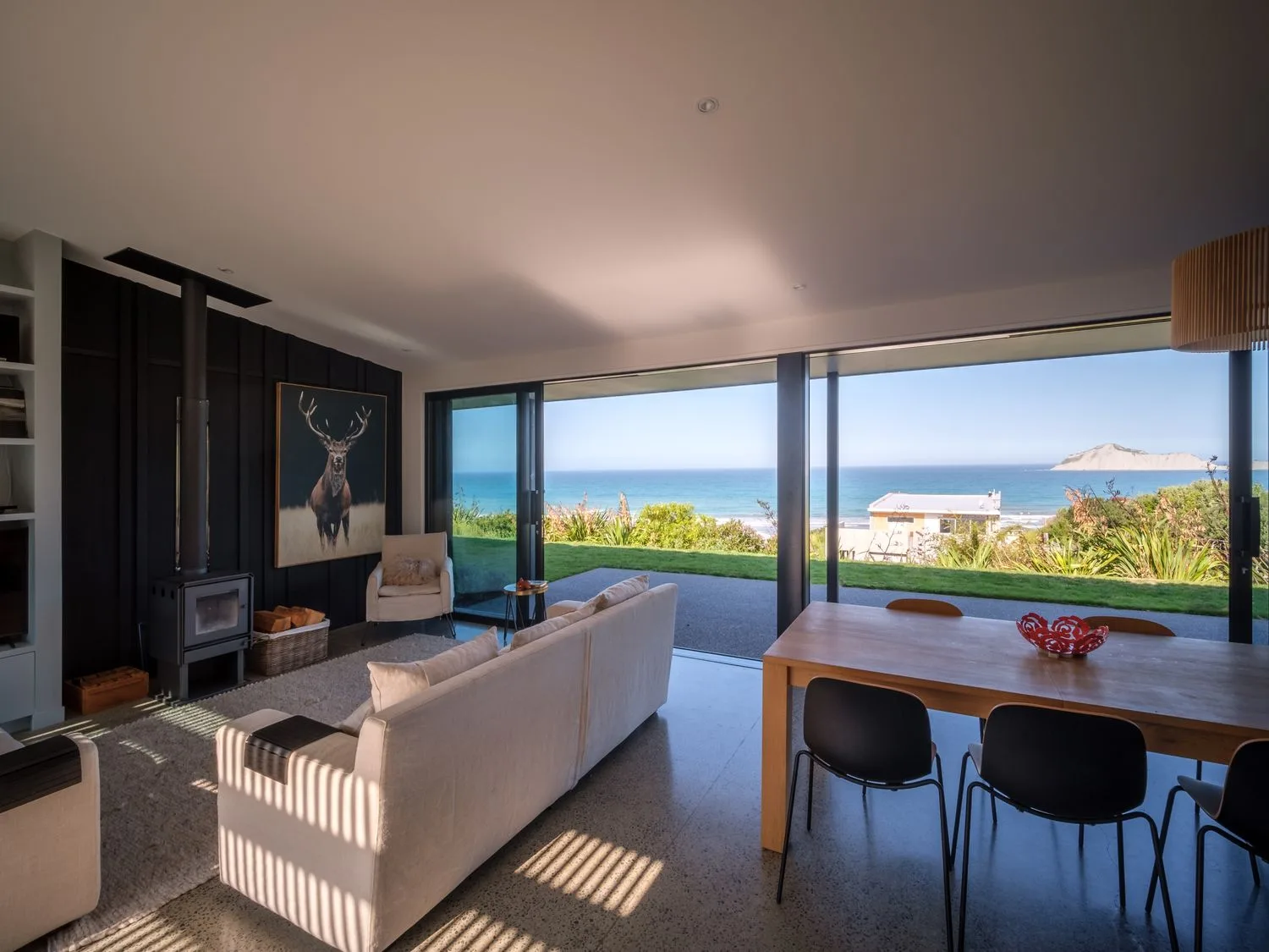 Una sala de estar con una estufa de leña, un gran cuadro de un ciervo y una mesa de comedor con sillas orientadas hacia las vistas al océano en Lightning Ridge Views Waimarama Holiday Home, en Waimarama